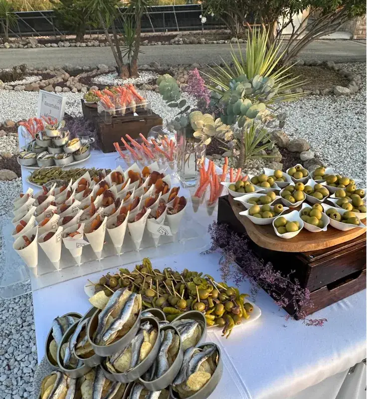 Mesa de catering de El Hogar montada al aire libre con conos de embutido, encurtidos y aceitunas